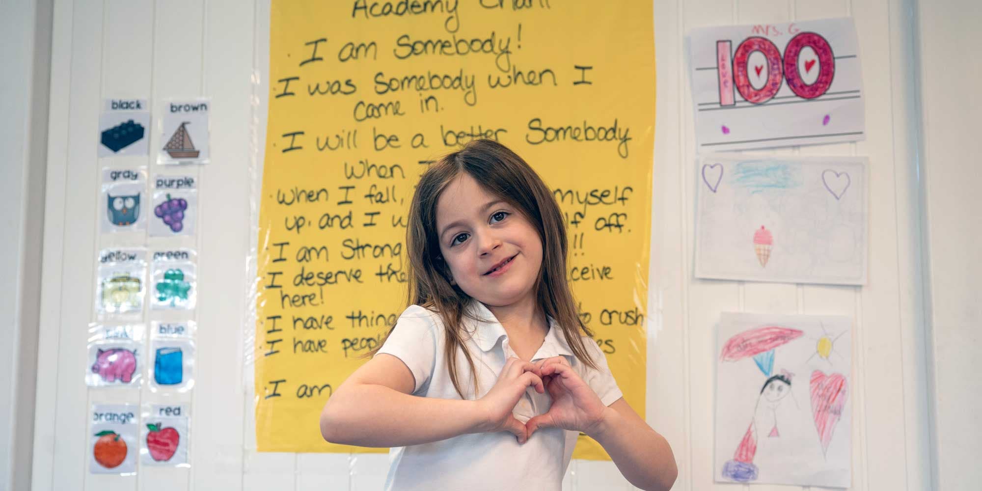 Smiling student making a heart shape with their hands in a classroom.