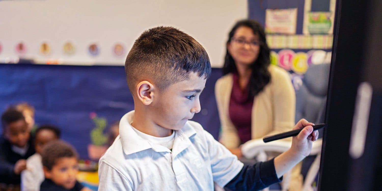 Student using a stylus on an interactive display as a teacher looks on.