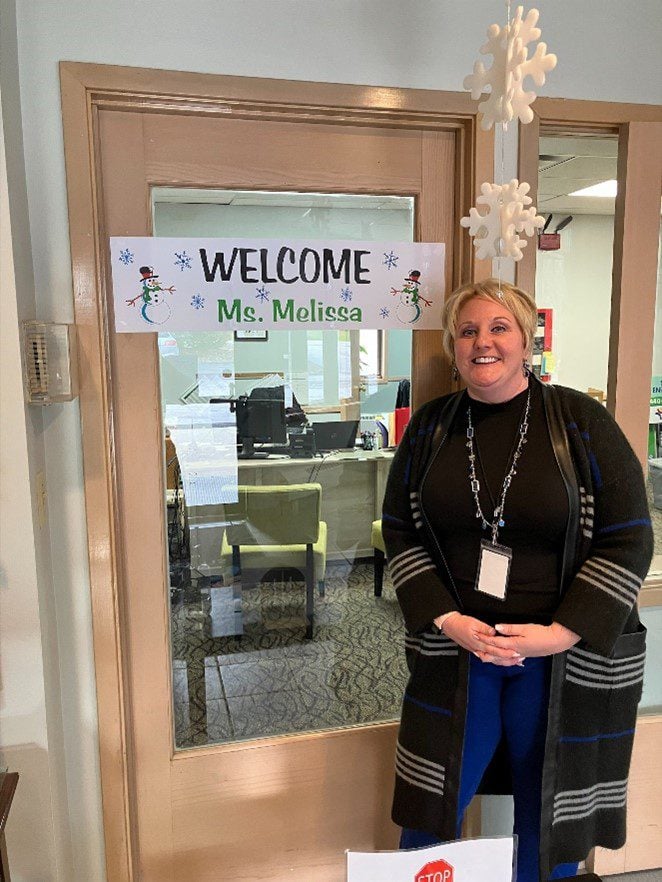 Adult smiling by a door with a "Welcome Ms. Melissa" sign.